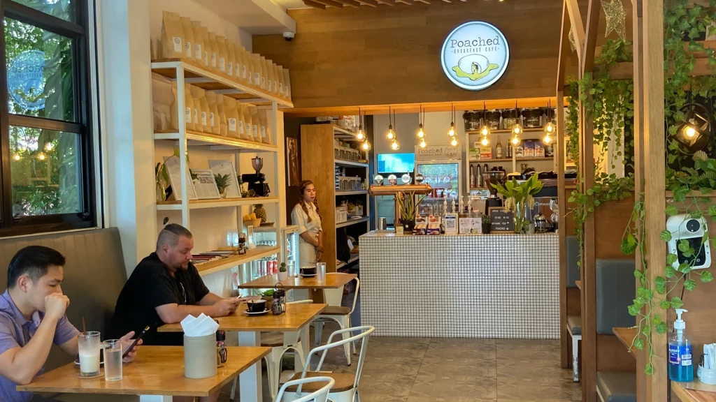 A view of the interior of Poached Breakfast Cafe, Phuket, Thailand. On the right, there are booths decorate with foliage, on the left there are wooden tables and chairs. 2 Customers sit with their food. There are shelves of coffee beans and nordic decor and at the back in the centre is a coffee bar with the espresso machine, wine on tap, a large sign and hanging bulb lights. One of the best cafes for digital nomads in Phuket.