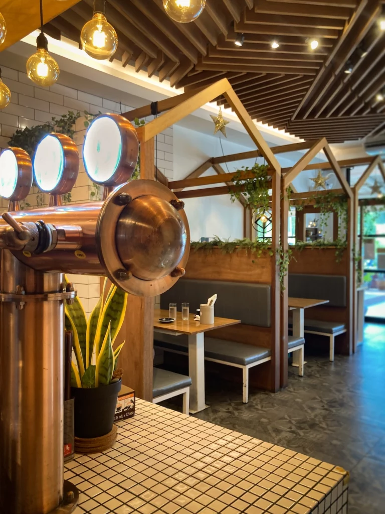 The view from the bar at Poached Breakfast Cafe, Chalong, Phuket, Thailand. Copper coloured wine on tap, under hanging lights take up the foreground. To the side, there are 2 wooden booths. This is one of the best cafes for digital nomads in Phuket.