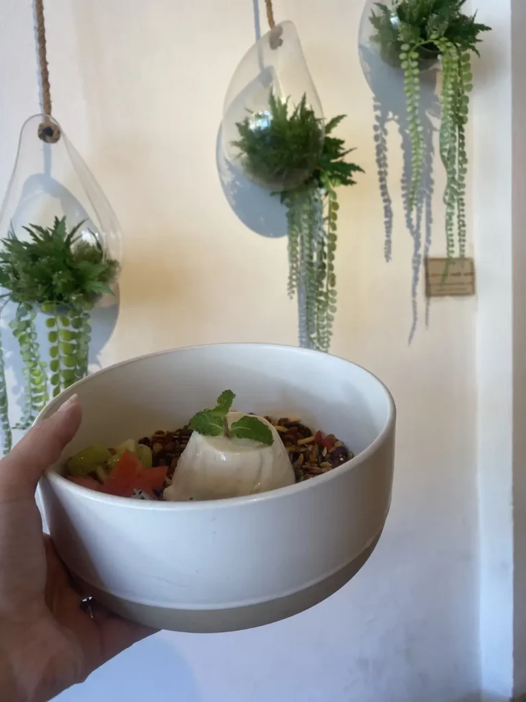 Someone holding up a bowl of granola topped with a beautiful dome of breakfast panna cotta with fresh seasonal fruit and a mint leaf. Only at the spot with the best juice near Chalong - Poached Breakfast Cafe, Chalong, Phuket, Thailand