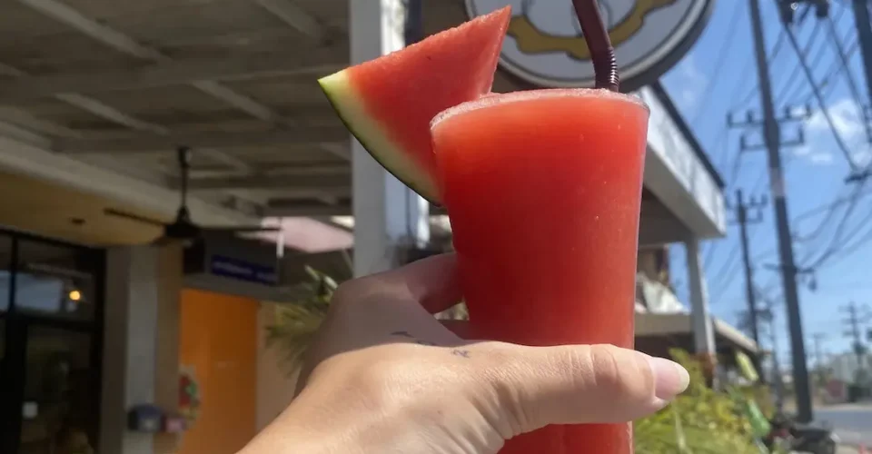 Watermelon smoothie in the sunshine being held up to our sign by a hand. A great alternative to street food in Chalong. Only at Poached Breakfast Cafe, Chalong, Phuket, Thailand